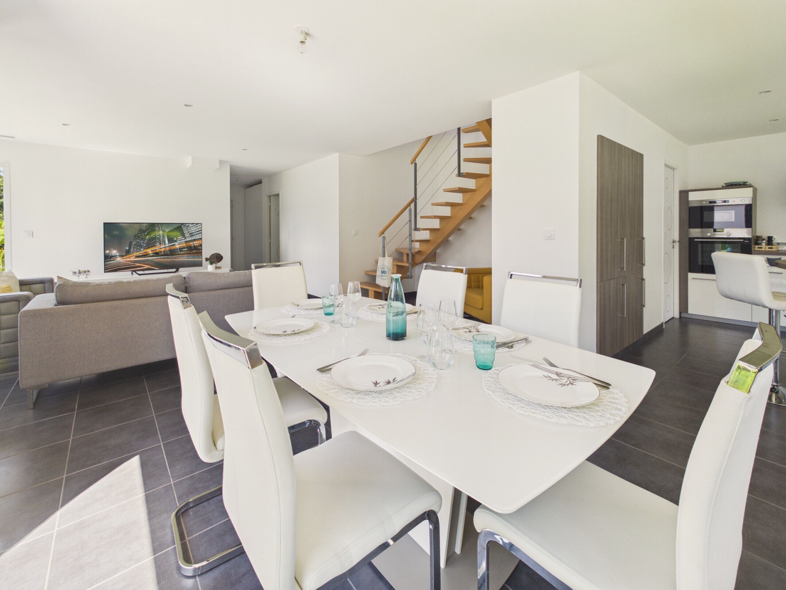 A modern dining area with a white table set for four, a staircase, and a kitchen in the background.