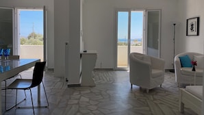 Une pièce comprenant une table à manger, des chaises, un canapé blanc, un lampadaire et une vue sur l’extérieur, accessible par une porte coulissante.