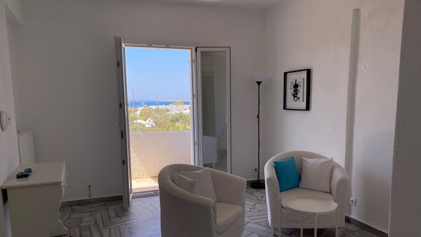 Une pièce avec un canapé blanc, une petite table, une chaise et un lampadaire. Une porte donne acc ès à un balcon extérieur d’où l’on peut admirer la mer.