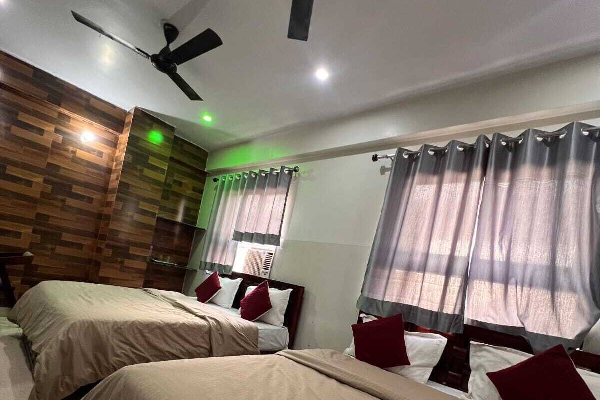 A hotel room with two beds, a ceiling fan, a wooden accent wall, and a window with curtains.