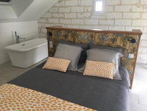 Un dormitorio con cama, dos almohadas, cabecera con estampado tropical, bañera y techo inclinado.