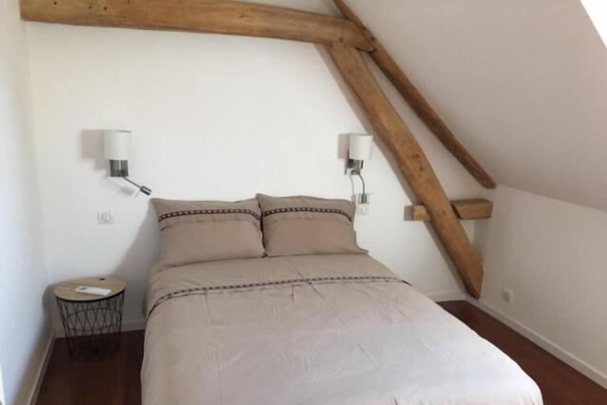 A bedroom with a bed, two wall-mounted lamps, a round nightstand, and exposed wooden beams.