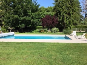 A rectangular swimming pool with a grassy lawn, trees, and two white lounge chairs.