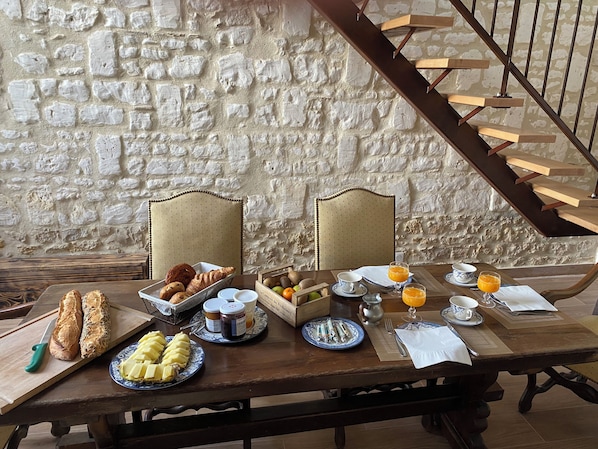 A breakfast setting with bread, pastries, fruits, and beverages on a wooden table against a stone wall.
