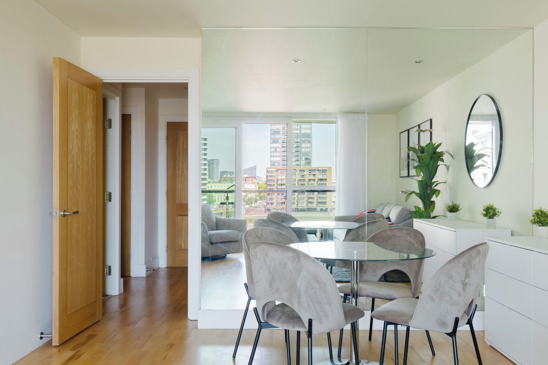 Un espace de repas moderne avec une table et des chaises en verre, un canapé et une vue sur des immeubles de grande hauteur.