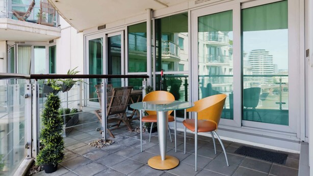 Ein Balkon mit Tisch und Stühlen, einem Blumentopf und Blick auf die Stadt durch große Fenster.