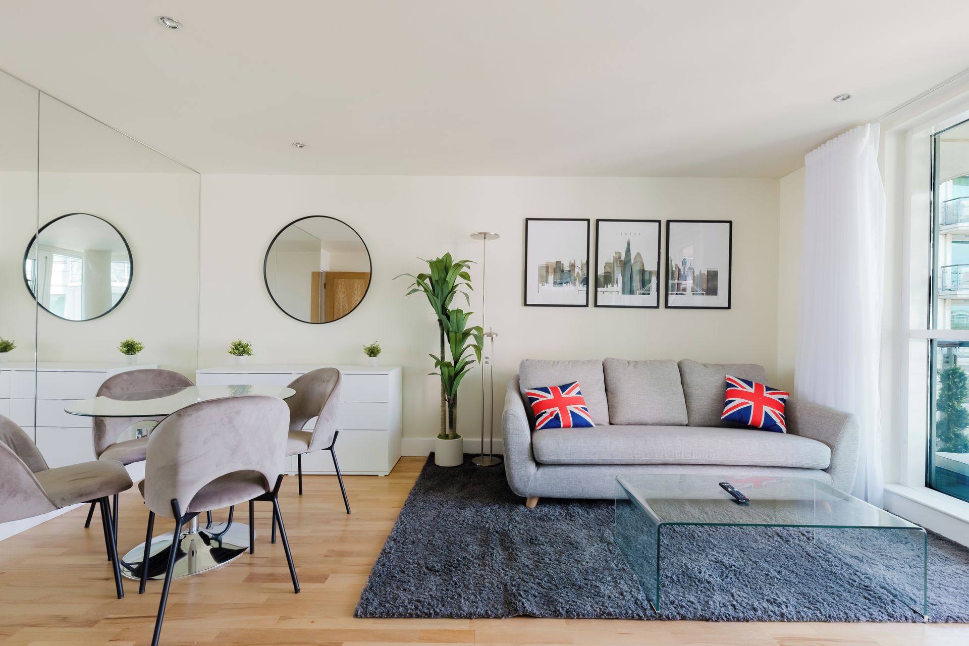 A modern living room with a grey sofa, two chairs, a round mirror, a dining table, and a glass coffee table. There are three framed pictures on the wall and a large window with curtains.