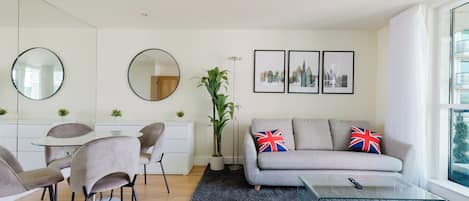 A modern living room with a grey sofa, two chairs, a round mirror, a dining table, and a glass coffee table. There are three framed pictures on the wall and a large window with curtains.