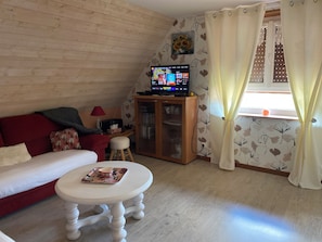 A cozy living room with a wooden wall, a red sofa, a white coffee table, a TV on a wooden cabinet, and a window with curtains.