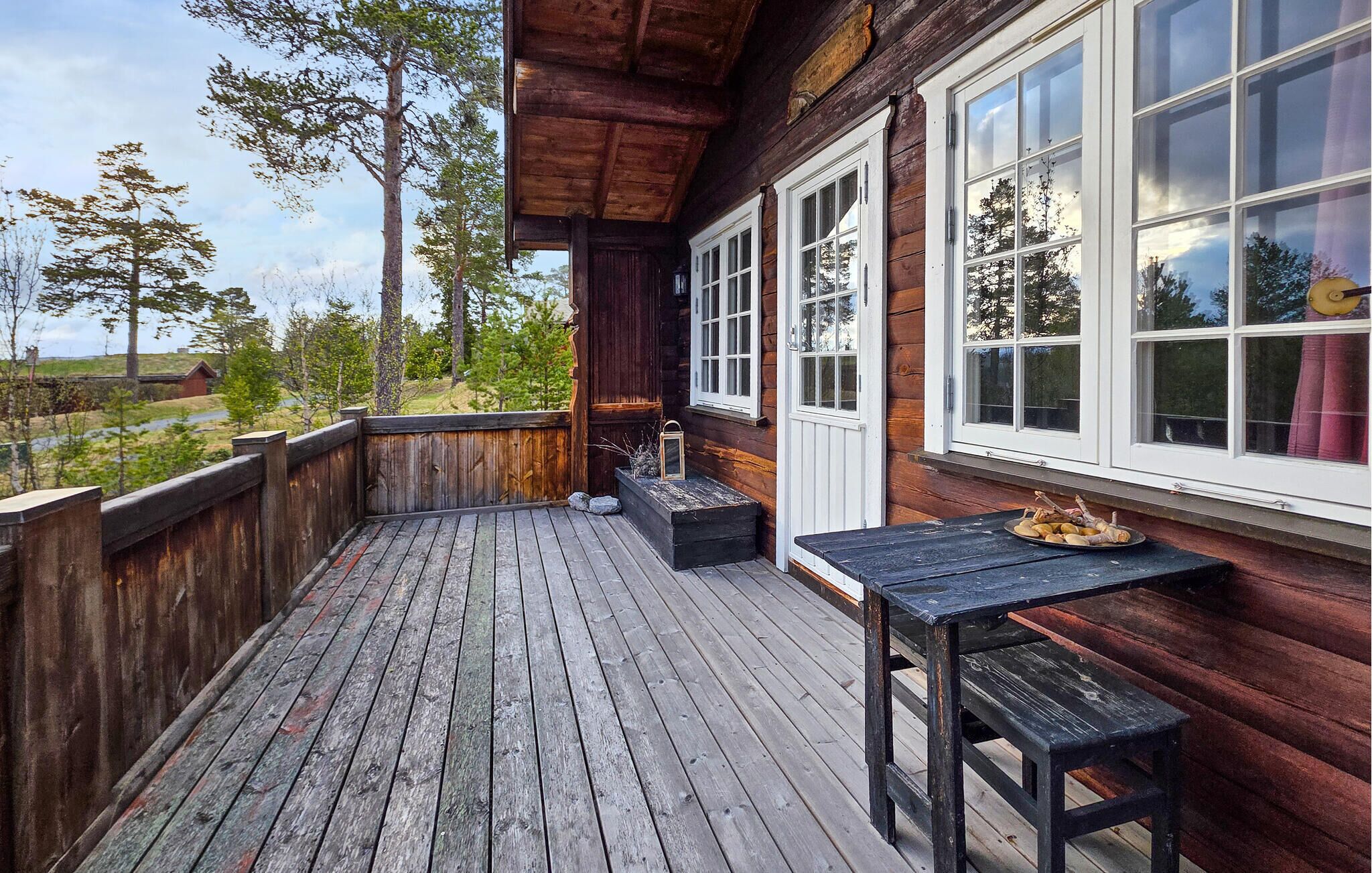 A wooden deck with a table and benches, a window with white frames, and a view of trees and a distant building.