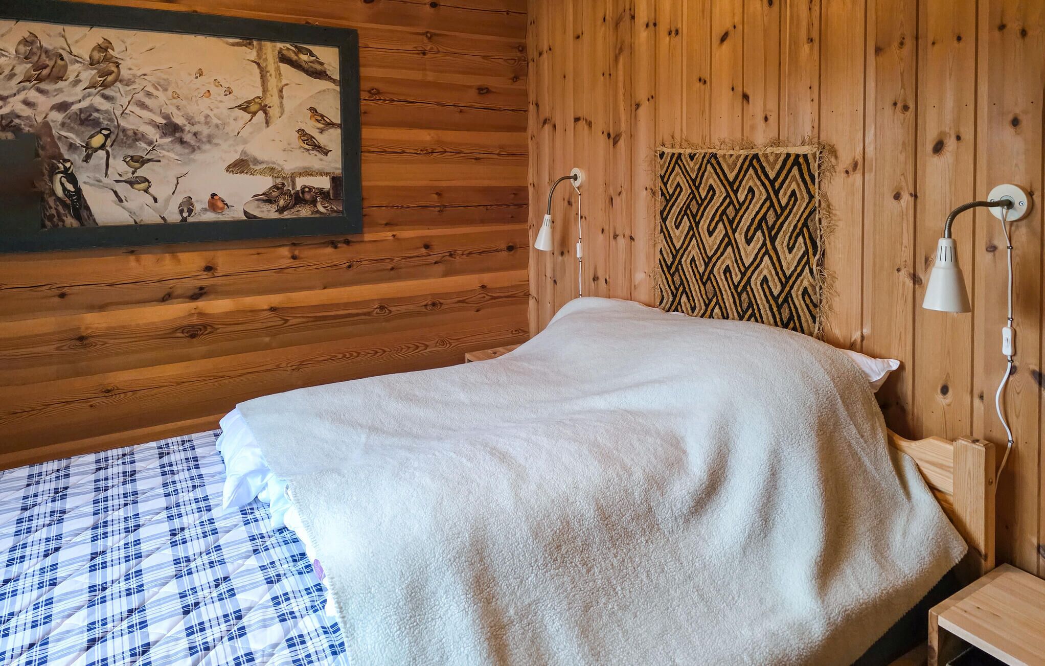 A wooden cabin room with a bed, a wall-mounted lamp, and a painting of birds.