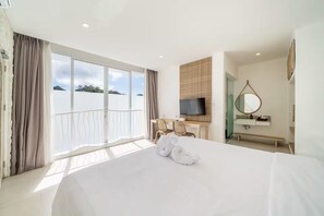 Une chambre d’hôtel avec un grand lit, un bureau avec des chaises, une télévision, une salle de bain avec un miroir et une vue sur la mer.
