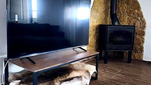 A living room with a large flat-screen TV on a wooden stand, a stone wall, and a wood-burning stove.