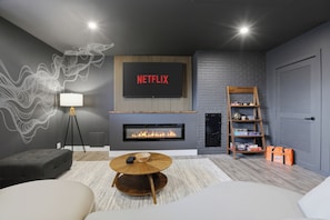 A modern living room with a fireplace, a Netflix sign, a bookshelf, and a cozy seating area. - NEW! Designer Home - King Beds, Glass Sunroom, Game Room, BBQ (Southfield)