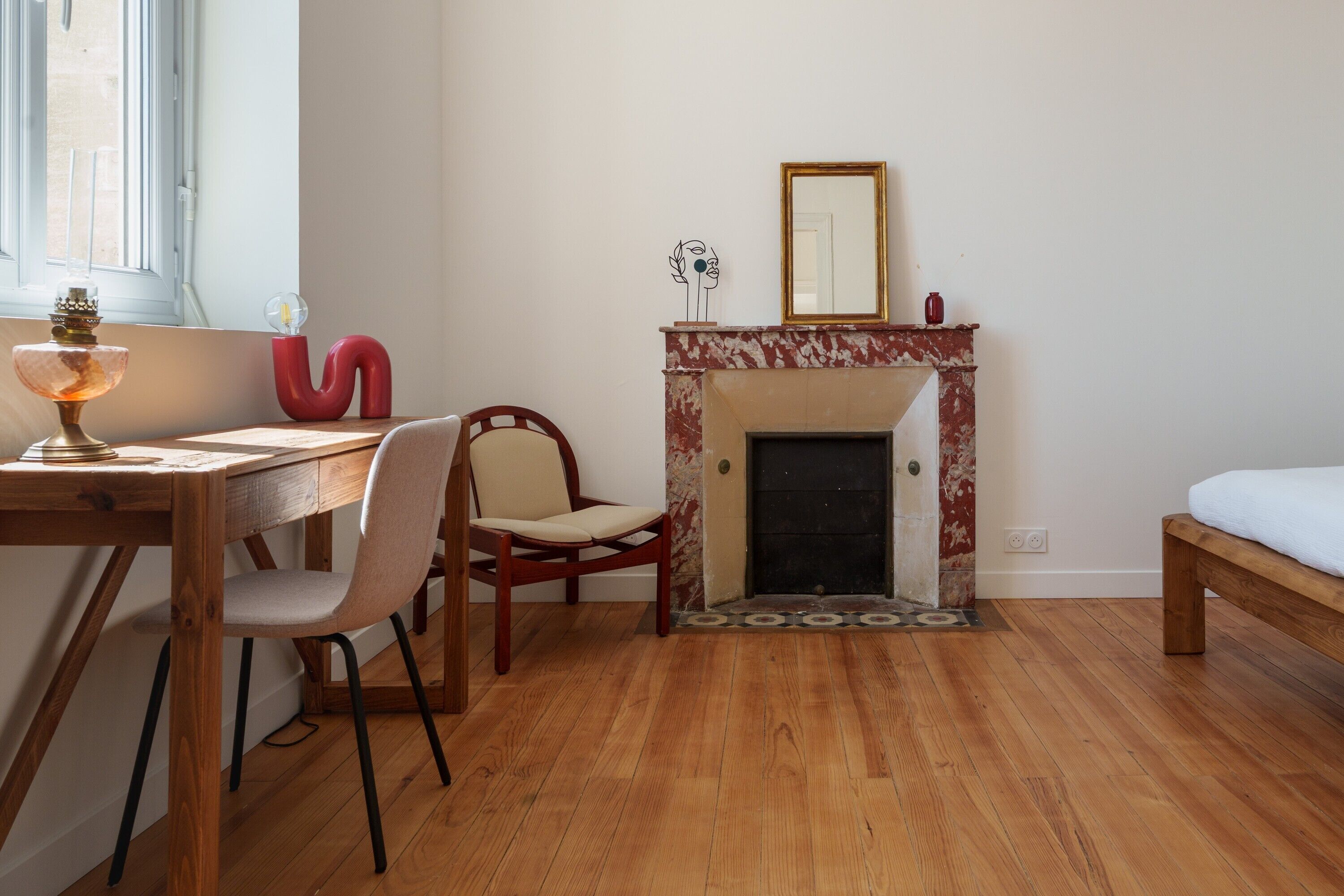 Una habitación con piso de madera, chimenea con un espejo encima, una silla, una mesa con una lámpara y una cama.
