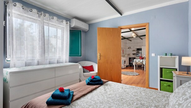 Une chambre à coucher avec un lit, une fenêtre avec des rideaux, une chaise, un bureau et, au fond, une petite cuisine.
