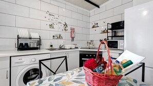 Une cuisine avec des carreaux blancs, un lave-linge, un four à micro-ondes et une table sur laquelle se trouve un panier contenant une rose et une bouteille.