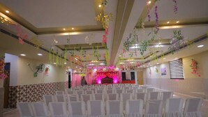 Une salle de banquet avec des rangées de chaises blanches, une scène décorée de rose et de rouge, et des plantes suspendues.
