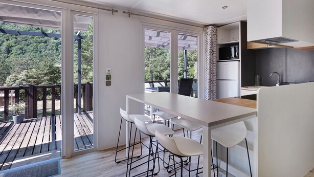 Eine moderne Küche mit Holzterrasse, weißen Barhocker und Blick auf ein bewaldetes Gebiet.
