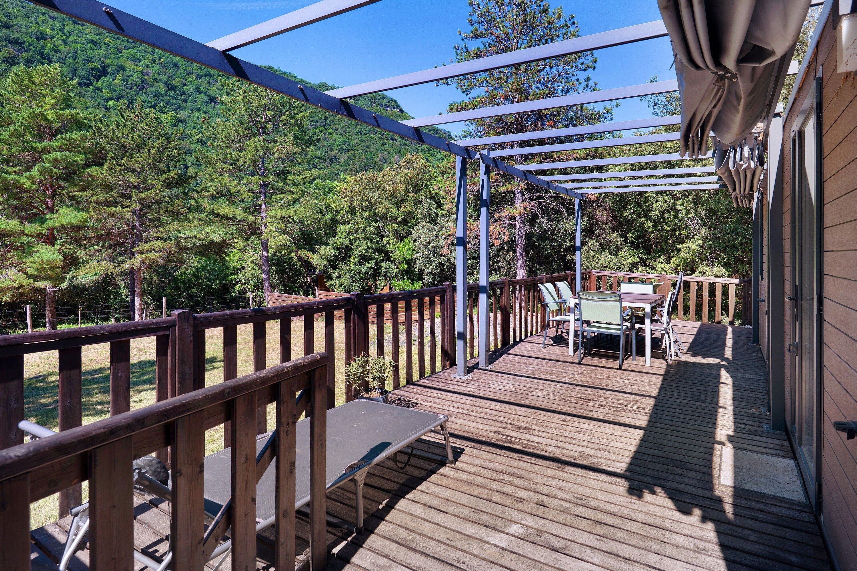 Une terrasse en bois agrémentée d’une pergola, de mobilier d’extérieur et offrant une vue sur une colline boisée.