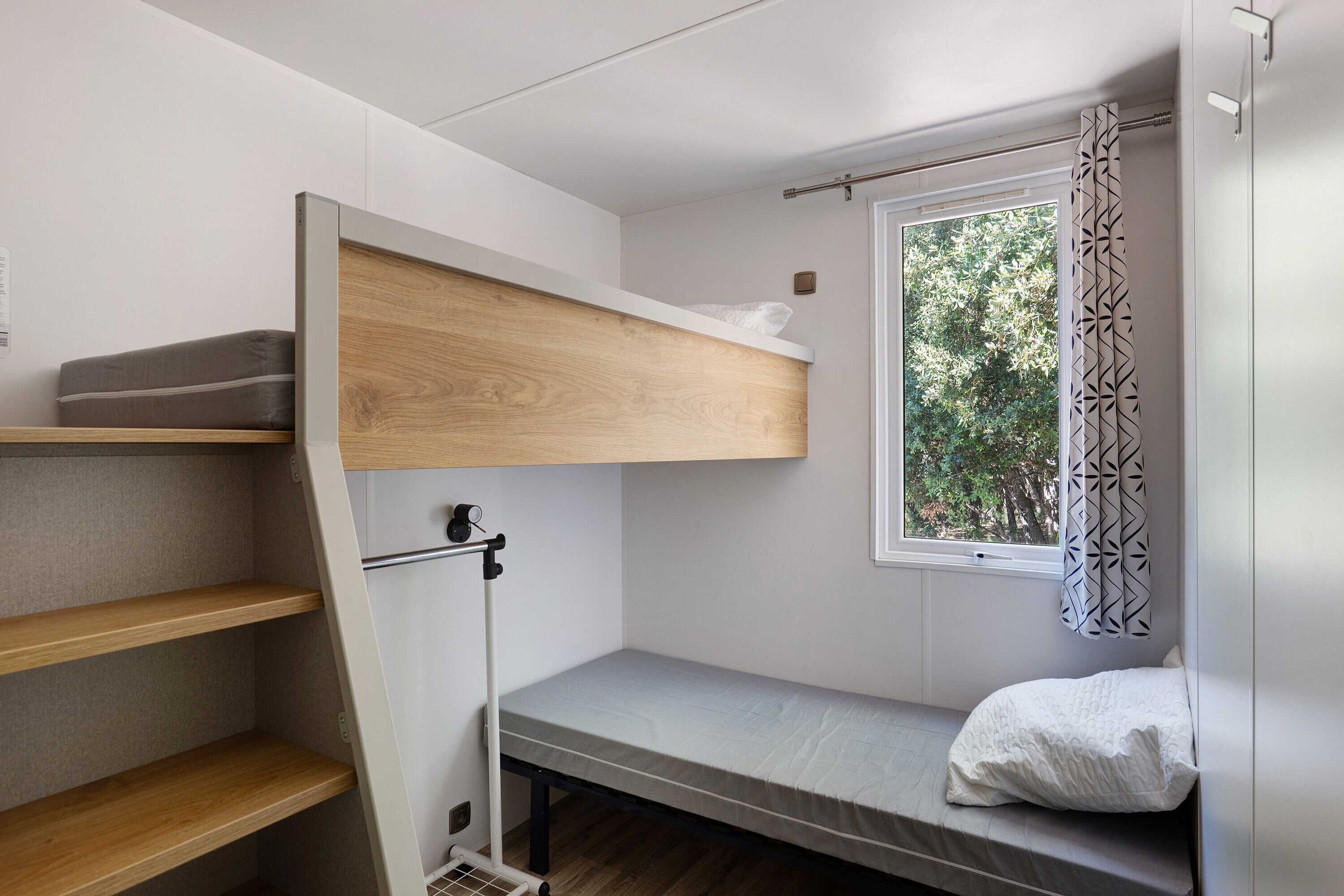 Un lit superposé avec une étagère en bois, une fenêtre ornée d’un rideau à motifs et un oreiller blanc.