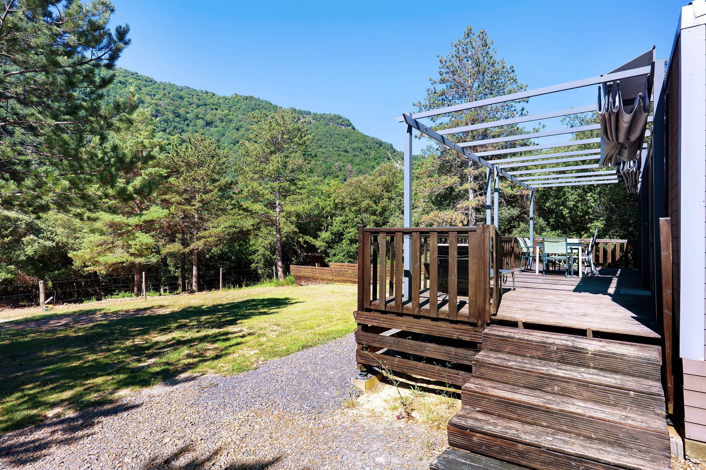 Una terraza de madera que conduce a un edificio con una pérgola, rodeado de un césped y una colina boscosa.