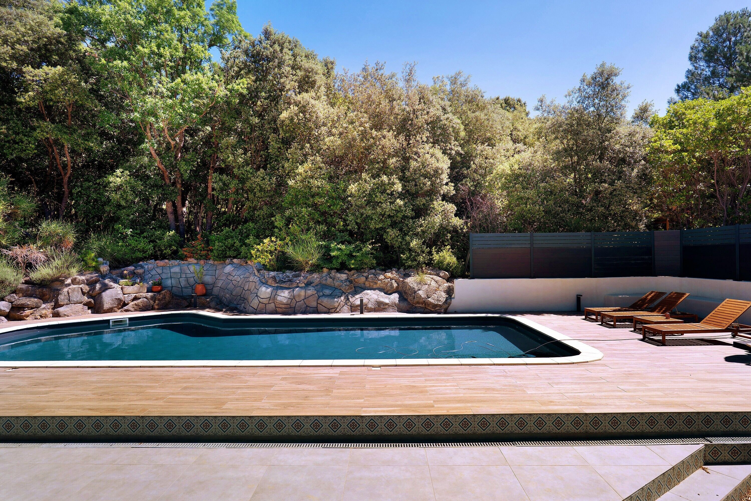 Une piscine avec une terrasse en bois, des chaises longues et un mur en pierre agrémenté de plantes.