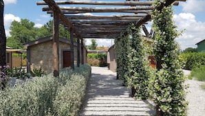 A rustic stone building with a wooden pergola over a gravel path, surrounded by greenery and flowering plants. - Agriturismo Resort Il Foionco (Massa Marittima)
