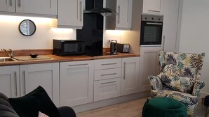 Una cocina moderna con armarios blancos, microondas, tostadora y horno. En la zona de estar, hay un sofá oscuro y un sillón estampado.
