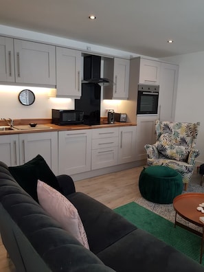 A modern kitchen with white cabinets, a microwave, toaster, and oven. There is a dark sofa and a patterned armchair in the living area. - Chic, modern, comfortable 2-bed apt directly opposite beach in town centre (Portrush)