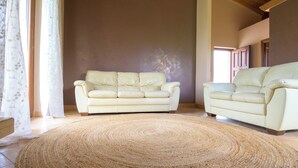 Een woonkamer met twee beige banken, een geweven vloerkleed en een groot raam met dunne gordijnen.