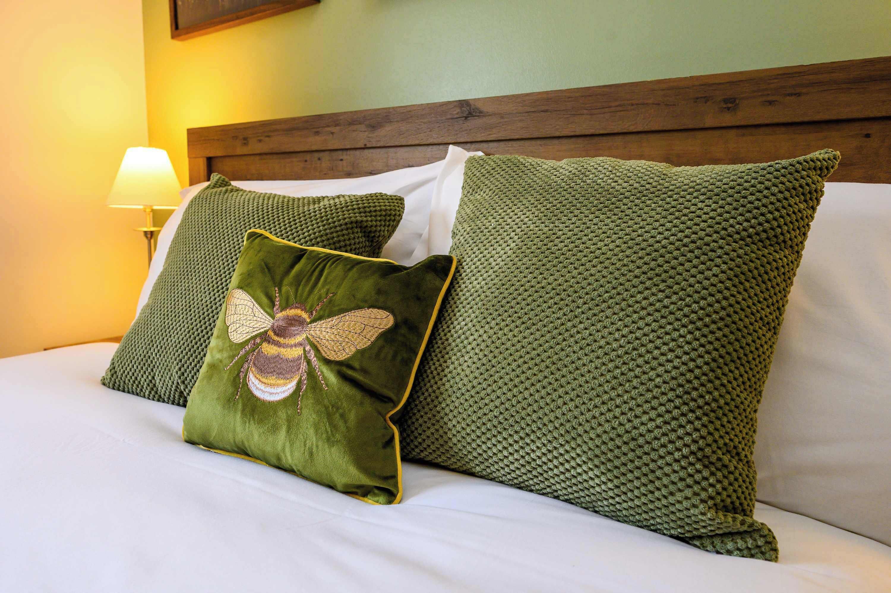Ein Bett mit grünen Kissen und einem mit Bienen bestickten Kissen, ein hölzernes Kopfteil und eine Lampe auf dem Nachttisch.