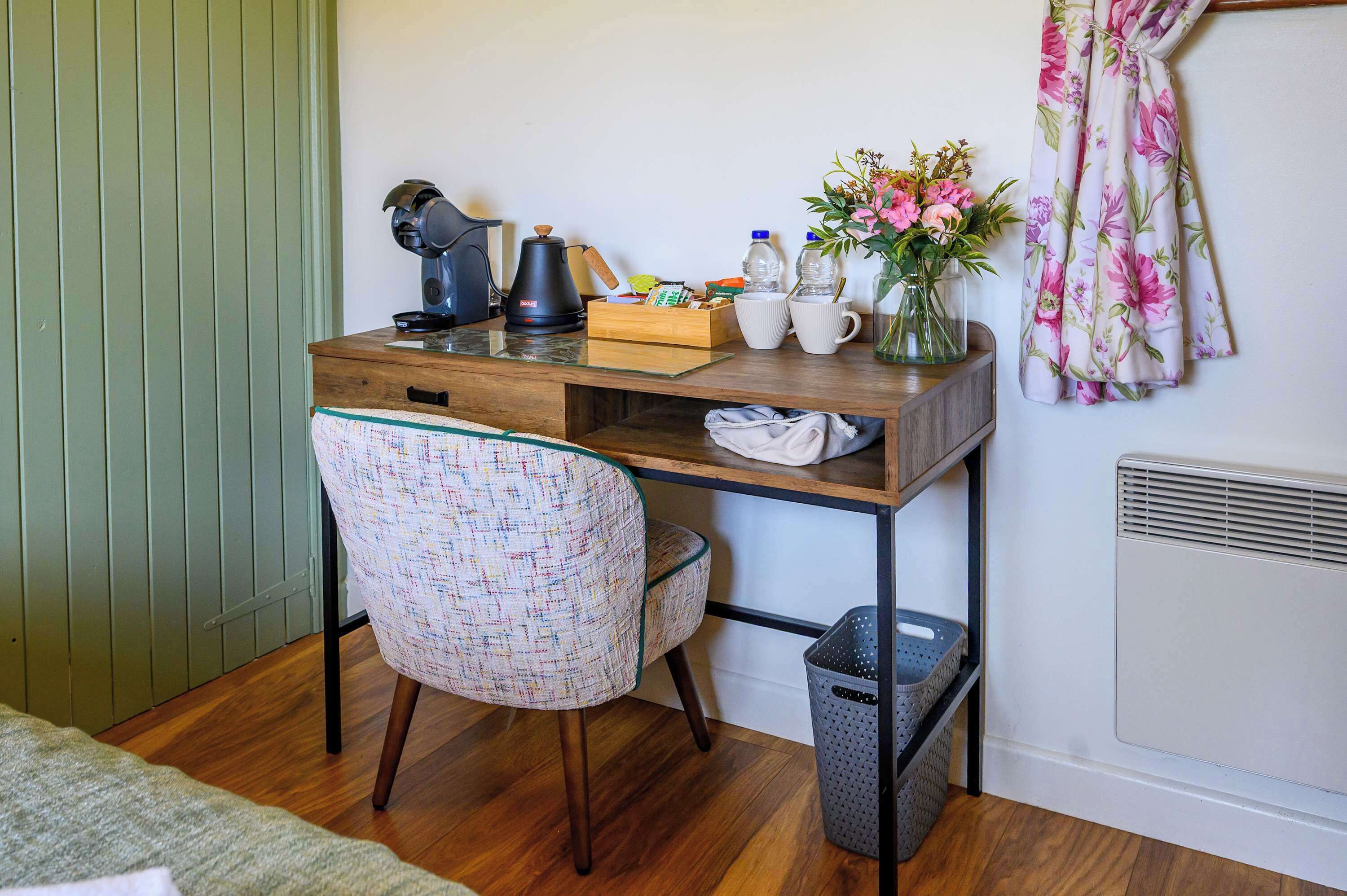A wooden desk with a chair, a kettle, cups, a vase with flowers, and a trash bin.