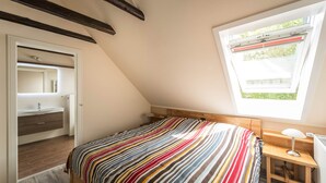 A bedroom with a sloped ceiling, a window with a view of greenery, a bed with a striped bedspread, a bedside table with a lamp, and a wooden headboard. - Harleblick Carolinensiel (Wittmund)