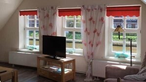 A room with a sloped ceiling, a television on a wooden stand, a window with red valance, and a radiator. - Harleblick Carolinensiel (Wittmund)
