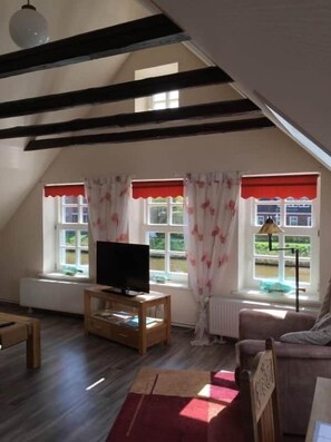 Ein Zimmer mit schrägem Dach, einem Fernseher auf einem Holzboden, einem Fenster mit rotem Vorhang und einem Heizkörper.