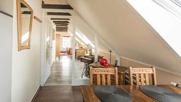 A loft-style room with a sloped ceiling, wooden beams, a dining area with a table and chairs, and a view of the living area through an open doorway. - Harleblick Carolinensiel (Wittmund)