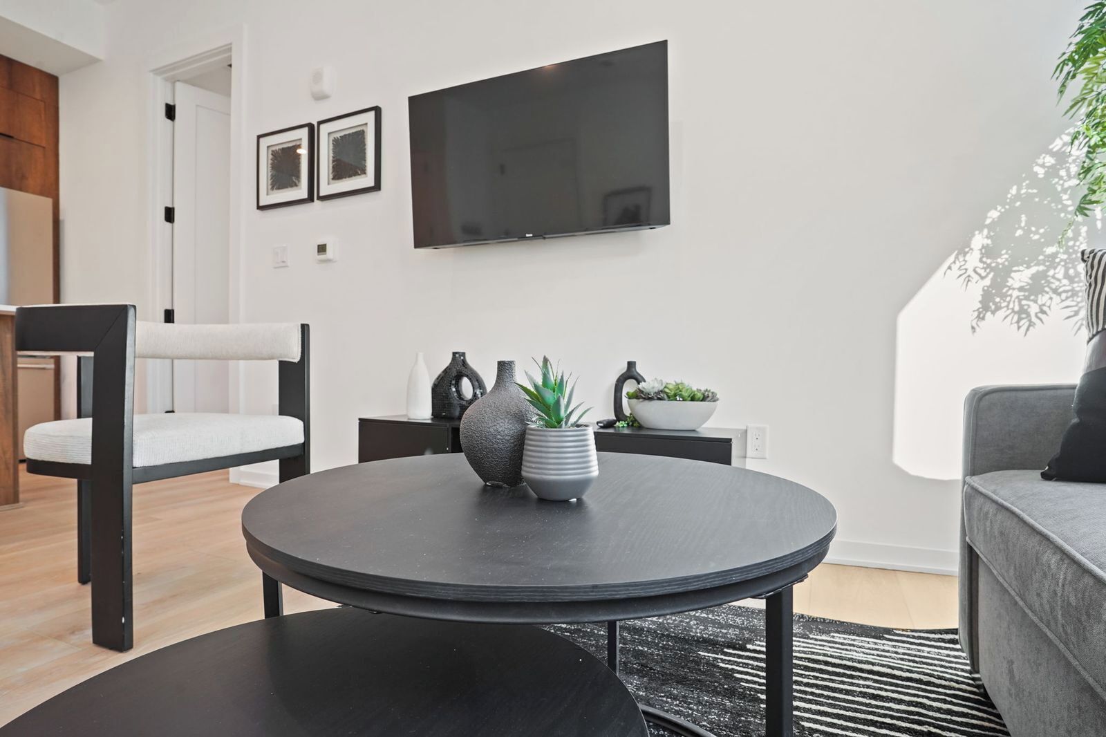A modern living room with a round black table, a grey sofa, a TV mounted on the wall, and a potted plant.
