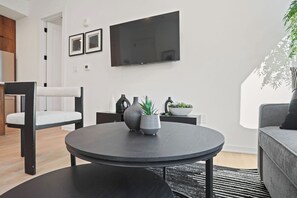 Una sala de estar moderna con una mesa redonda negra, un sofá gris, un televisor montado en la pared y una planta en maceta.