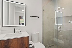 Un baño moderno con un espejo grande, un lavamanos blanco, un mueble de tocador de madera, un inodoro y una cabina de ducha con paredes de vidrio.