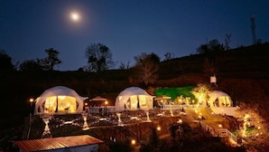 Three dome-shaped structures with internal lighting, surrounded by a stone wall and a wooden fence, set against a night sky with a visible moon. - Manzil Escapes (Dhanaulti)