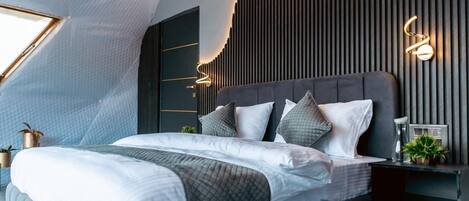 A modern bedroom with a sloped ceiling, a bed with multiple pillows, a bedside table with a lamp, and a wall-mounted light fixture.