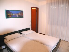 Ein Doppelbett mit weißen Leinen, ein Holzkopfende, ein Nachttisch, ein gerahmtes Stadtbild und ein durchsichtiger Vorhang.