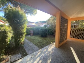 Ein überdachter Außenbereich mit einem Betonweg, der zu einem Rasenplatz führt, umgeben von Bäumen und einem Zaun.