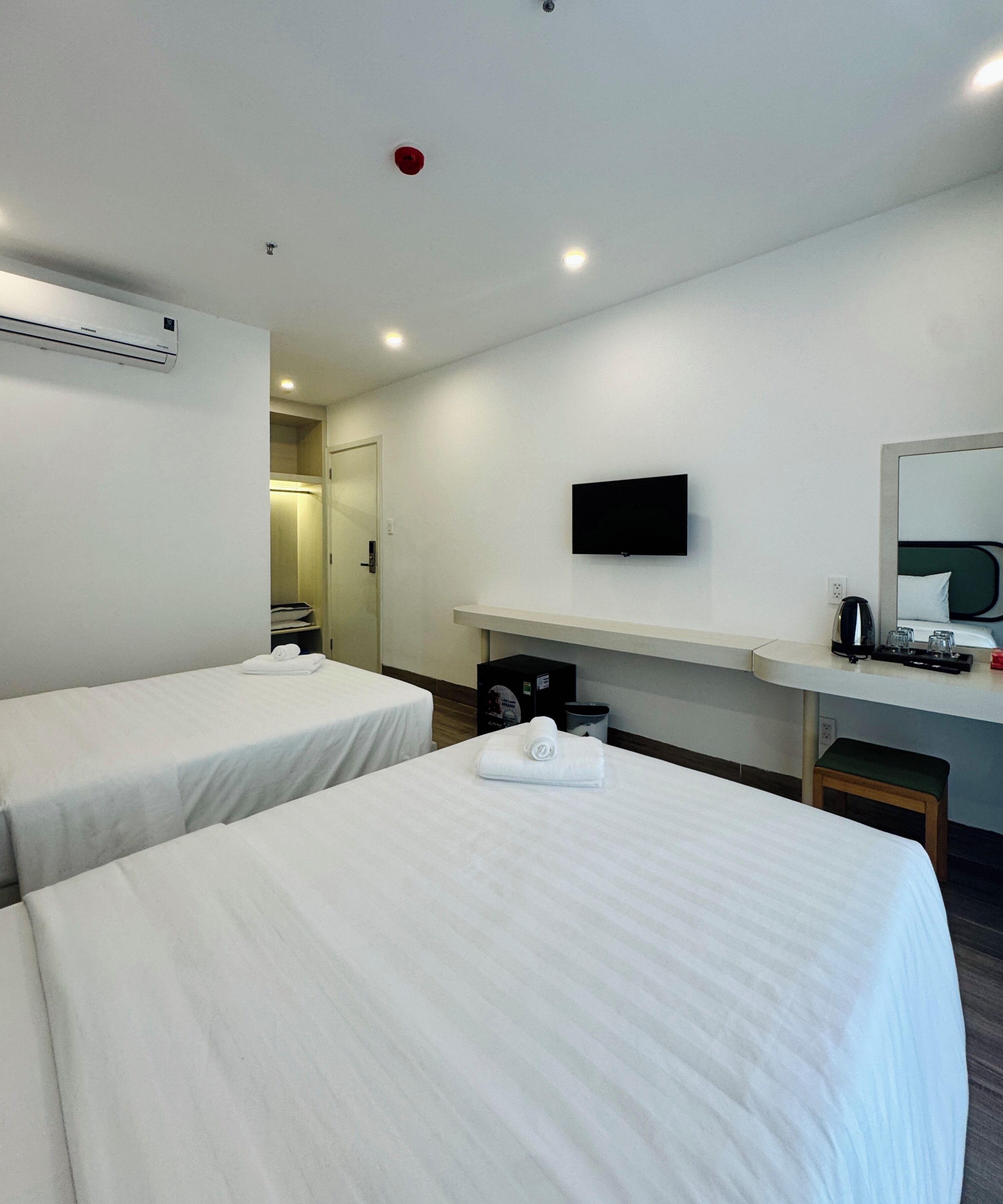 A hotel room with two beds, a desk with a flat-screen TV, a mirror, a kettle, and a trash bin.