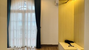 Hotelkamer met houten vloer, een bed met matras en kussens, een nachtkastje met lamp, een raam met dunne gordijnen en een wandgemonteerde airconditioning.