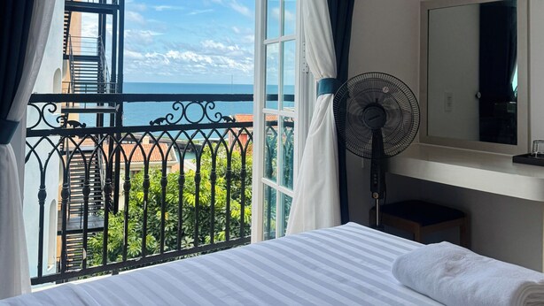 Een hotelkamer met een bed, een ventilator, een spiegel en uitzicht op de zee door een groot raam.