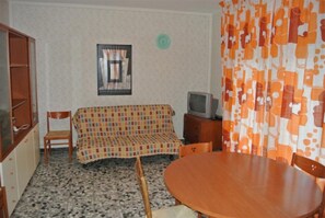 Ein Zimmer mit einer gemusterten Couch, einem Fernseher auf einem Holzschrank, einem runden Holztisch und einem gemusterten Vorhang.