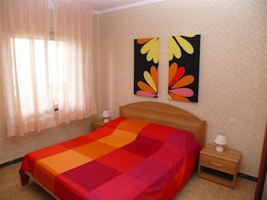Ein Schlafzimmer mit einem Bett, zwei Nachttischen, einem Fenster mit Vorhängen und zwei an der Wand befestigten Kunstwerken.