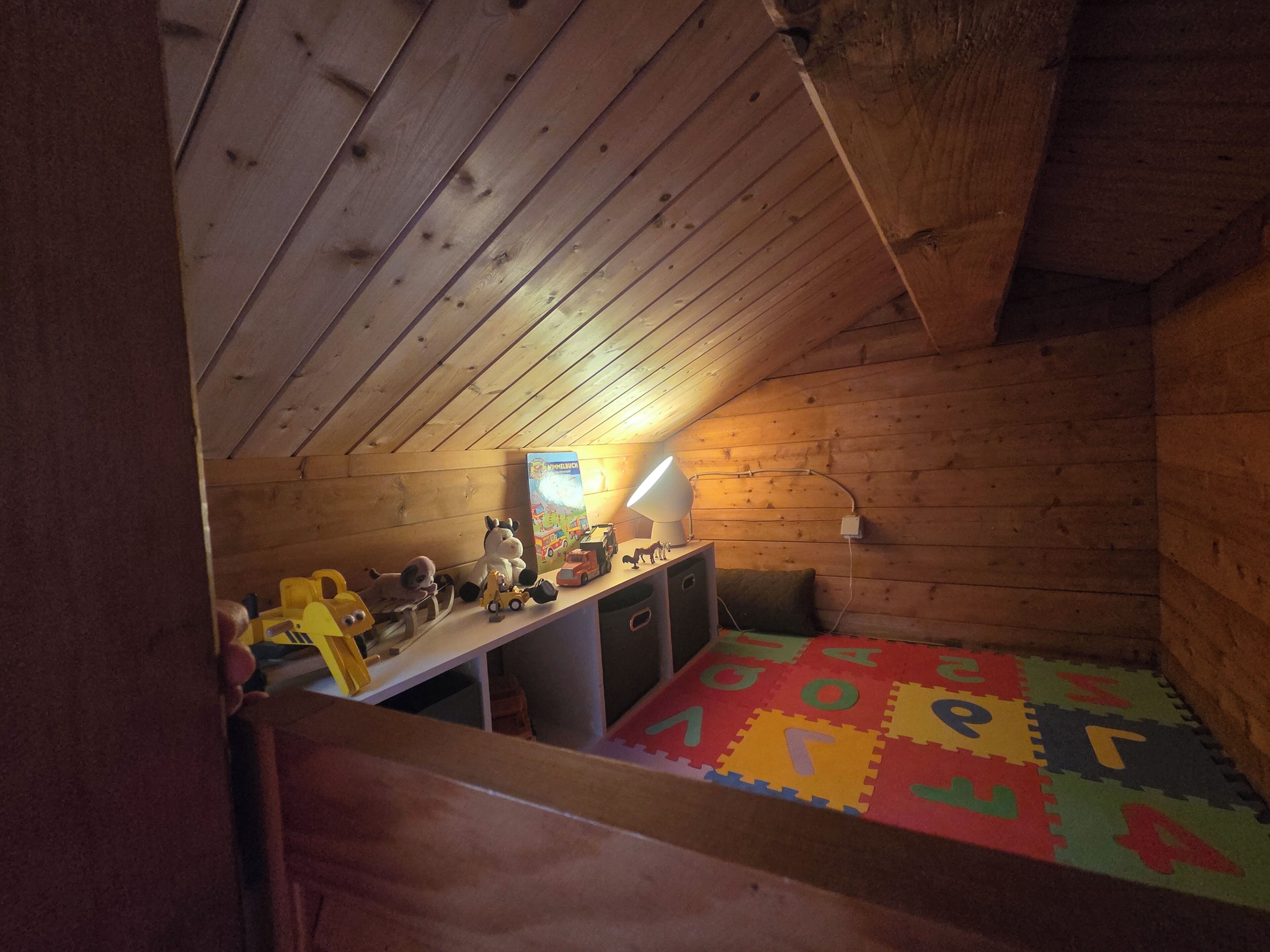 A room with a sloped ceiling, wooden walls, a desk with a lamp, a bed with a colorful mat, and toys.